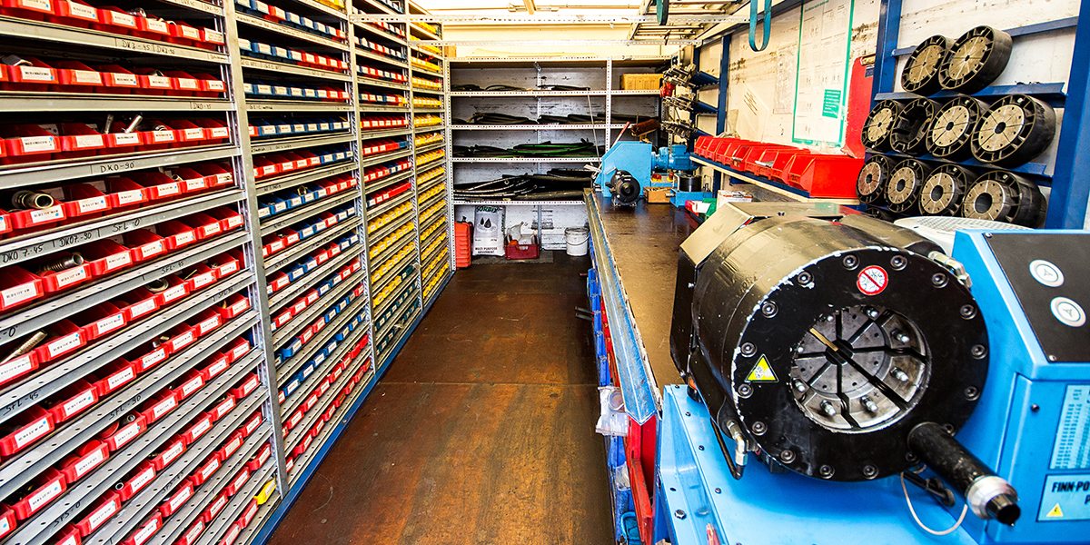 Innenraum des Presswagens: Regale mit Ersatzteilen in roten und blauen Kästen, Schlauchrollen und eine Schlauchpresse an der Werkbank