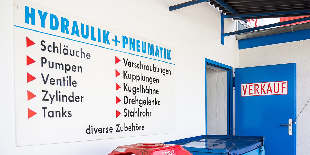 Schild am Ladengeschäft „Hydraulik + Pneumatik“: Schläuche, Pumpen, Ventile, Zylinder, Tanks, Verschraubungen, Kupplungen, Kugelhähne, Drehgelenke, Stahlrohr und diverse Zubehöre, daneben die blaue Verkaufstür